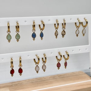 Op de afbeelding: Een collectie goudkleurige oorringen met bungelende druppelvormige bedels in diverse kleuren, waaronder lichtgroen, blauw, grijs, wit, rood en roze. Elke oorbel heeft een klein decoratief kraaltje aan de onderkant van de druppelvormige bedel.