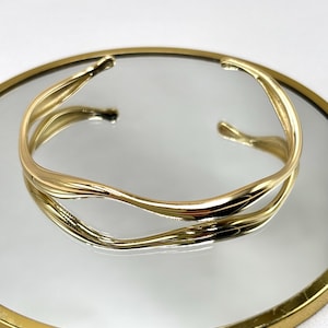 Könnte beinhalten: Ein goldfarbenes Armreif-Armband mit fließendem, organischem Design. Das offene Armband hat eine glatte, polierte Oberfläche und spiegelt sich im darunter liegenden Spiegel. Das Armband wird auf einem runden Spiegel mit goldfarbenem Rahmen präsentiert.