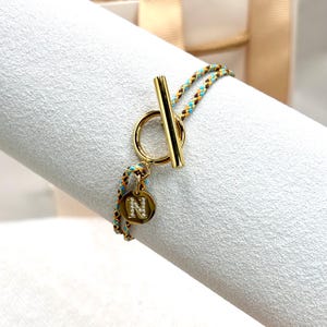 May include: Gold-coloured bracelet featuring a toggle clasp. The braided cord is in shades of blue, orange, and brown. A circular charm with the letter "N" is embellished with small, clear stones. The bracelet is displayed on a white surface.