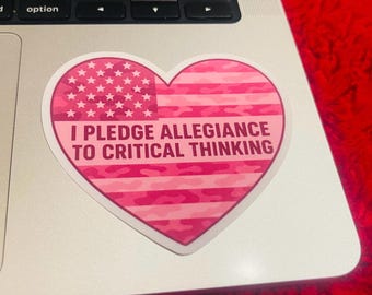 May include: A heart-shaped sticker with a pink American flag design. The sticker features the text "I PLEDGE ALLEGIANCE TO CRITICAL THINKING" in white. The sticker is on a silver laptop.