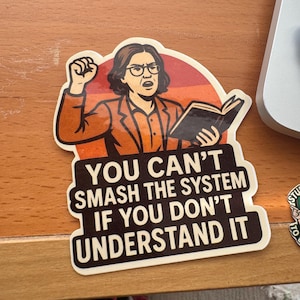 May include: A sticker featuring a cartoon illustration of a person holding a book and raising a fist. The sticker has the text "YOU CAN'T SMASH THE SYSTEM IF YOU DON'T UNDERSTAND IT" in white letters.