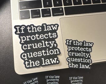 May include: Five black stickers with white text that reads, "If the law protects cruelty, question the law." The stickers vary in size and are arranged on a laptop keyboard. The text is in a bold, sans-serif font with a white outline around the letters.