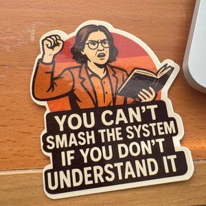 May include: A die-cut sticker featuring an illustration of a person with dark hair and glasses, wearing an orange blazer, holding a book, and raising a fist. The text reads: YOU CAN'T SMASH THE SYSTEM IF YOU DON'T UNDERSTAND IT, set against a red circle.