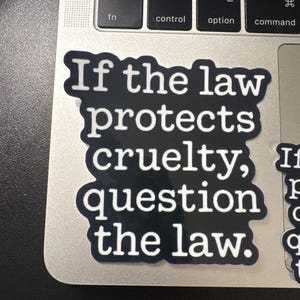 May include: A black sticker with white text that reads "If the law protects cruelty, question the law." The sticker is on a laptop keyboard. Additional smaller stickers with the same text are visible.