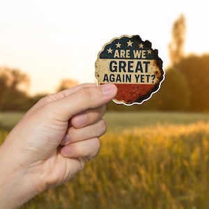 May include: A distressed, circular sticker with a patriotic design. The top section features a blue background with white stars, the middle section has white text that reads "ARE WE GREAT AGAIN YET?", and the bottom section is red.