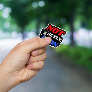 May include: A sticker with the words "NOT EXACTLY ON" in red, black, and blue. The sticker is held in a hand against a blurred background of trees and a path.