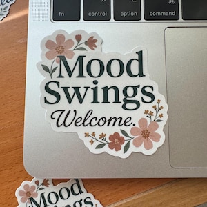 May include: Two stickers with floral designs and the text "Mood Swings Welcome." The stickers feature a white background with dark green lettering and pink and brown floral accents. One sticker is on a laptop.