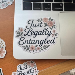 May include: A white sticker with the text "Just Legally Entangled" in a cursive font, surrounded by a floral wreath of pink roses and green leaves. The sticker is on a laptop, with other stickers visible.
