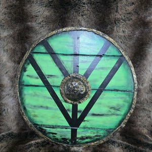 May include: A circular wooden shield featuring a vibrant green finish with horizontal wood grain texture. It is decorated with black painted lines forming a geometric pattern and a central metallic boss with a textured, weathered bronze appearance.