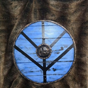 May include: A circular wooden shield featuring a light blue painted surface with black painted lines forming a tree-like pattern. The shield has a dark, textured metal rim and a central, raised, hammered metal boss with a dark, weathered finish.