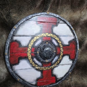 May include: A circular wooden shield featuring a white background with a prominent red cross design. The centre has a dark, textured dome surrounded by a gold-coloured ring with triangular patterns. The outer rim is dark grey with small gold studs.