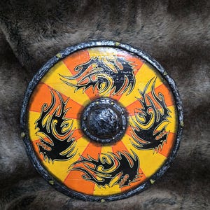 May include: A circular wooden shield featuring a vibrant sunburst pattern in alternating yellow and orange segments. Four stylised black dragon motifs are painted around a central dark, textured metal boss. The rim is finished in a dark, weathered metallic texture with small studs spaced evenly along the edge.