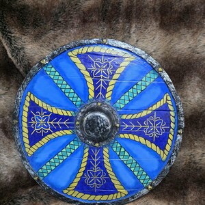 May include: A circular wooden shield featuring a vibrant blue surface with a painted yellow rope-like cross pattern. The centre has a dark, textured metal boss, and the outer rim is finished in a weathered silver tone with small gold studs.