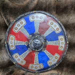 May include: A circular shield featuring a central metallic boss and a repeating pattern of eight silver-coloured hammers arranged radially. The background is divided into alternating blue and red segments, with a dark, textured metallic rim.