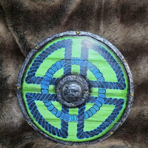 May include: A circular wooden shield featuring a bright green base with a painted blue and black rope-like knot pattern. The shield has a dark, textured metallic rim with small gold accents and a central raised metallic boss with a weathered finish.