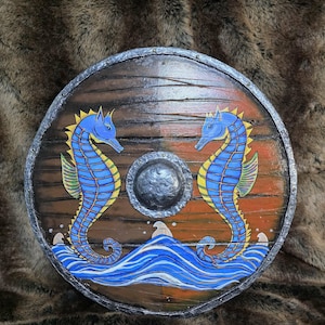 May include: A round wooden shield featuring a dark brown finish with a central metal boss. The surface is painted with two blue and yellow seahorses facing each other above stylised blue and white waves, all enclosed by a metallic silver rim.