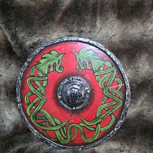 May include: A circular shield featuring a vibrant red background with two intertwined green serpent-like creatures painted on the surface. The shield has a dark, textured metallic rim and a central black dome, accented with small gold-coloured studs.