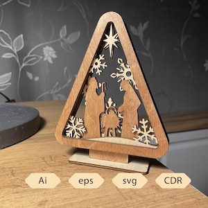 May include: A wooden Christmas tree-shaped decoration featuring a nativity scene with a star and snowflakes. The laser-cut design is made of light brown wood and stands on a small base. The bottom of the image shows the text "Ai", "eps", "svg", and "CDR".