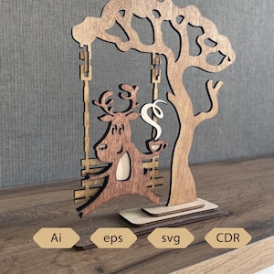 May include: Wooden decorative piece featuring a deer on a swing, holding a cup of coffee, under a tree. The design is laser-cut from wood. The base includes the text "Ai", "eps", "svg", and "CDR".
