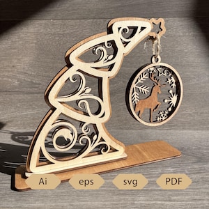 Puede incluir: Un expositor de madera con forma de árbol de Navidad con un diseño decorativo recortado. Un adorno redondo con un diseño de reno y copo de nieve cuelga de la parte superior. La base tiene las abreviaturas de tipo de archivo: Ai, eps, svg y PDF.