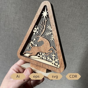 Peut inclure: Décoration de sapin de Noël en bois de forme triangulaire, avec un motif découpé d'un renne, de flocons de neige et d'une étoile. L'arbre est en bois brun clair et possède un socle. L'image comprend également le texte "Ai", "eps", "svg" et "CDR".