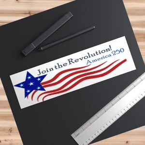May include: A white sticker with the text "Join the Revolution! America 250" in blue and red. The sticker features a blue star with white stars and red and white stripes, resembling the American flag. A ruler and two black pens are also visible.