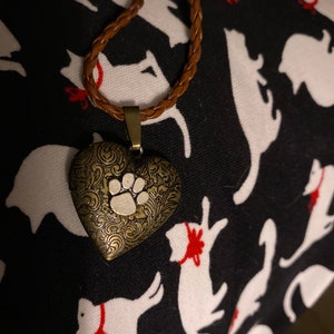 May include: A brown leather cord necklace with a gold heart-shaped pendant featuring a white paw print. The necklace is on a black fabric background with white silhouettes of dogs.
