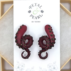 May include: A pair of deep red octopus tentacle earrings featuring dark, textured suction cups. The earrings are displayed on a white card printed with the text Petal & Pearl by Nidhi and a floral illustration at the bottom of the card.