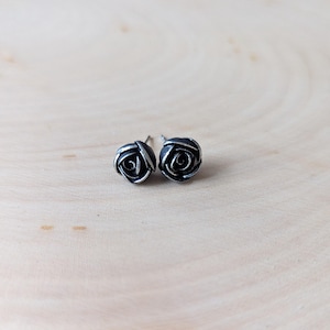May include: A pair of silver-toned rose stud earrings. Each earring features a detailed rose design with visible petals. The earrings are set against a light wooden background, showcasing the jewelry's intricate details.