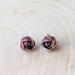 Può includere: Un paio di orecchini a bottone a forma di rosa. Gli orecchini sono di colore bordeaux scuro con bordi dorati. Gli orecchini sono montati su perni d'argento e sono esposti su una superficie di legno chiaro.