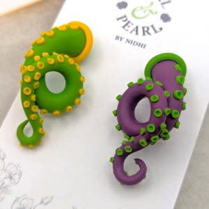 May include: A pair of stud earrings shaped like curled octopus tentacles. One tentacle is lime green with yellow suckers, and the other is purple with green suckers. They are displayed on a white card labeled Petal & Pearl by Nidhi.