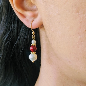 Può includere: Orecchino pendente color oro con una perla bianca, una perla rossa e una perla bianca più piccola. L'orecchino è sospeso a un gancio dorato e viene indossato sull'orecchio.
