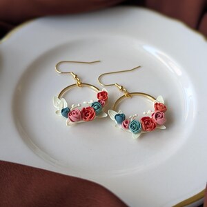 Può includere: Un paio di orecchini a cerchio dorati con motivi floreali. Ogni orecchino presenta piccoli fiori rossi, rosa e blu, accentuati da piccole foglie bianche. Gli orecchini sono esposti su un piatto bianco.