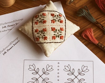 May include: A white fabric pincushion with a red and green embroidered floral pattern. The pincushion is on a white sheet of paper with a black and white floral pattern printed on it. The paper also has the text "Modernism&Vintage Folk Flowers Pattern" printed on it.