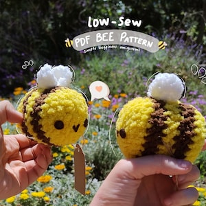 May include: Two handmade, plush bee amigurumi toys. The bees are yellow and brown with white wings. The text "low-sew PDF BEE PATTERN" is at the top. The bees have black eyes and are held in front of a blurred floral background.