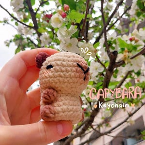 May include: A crocheted capybara keychain in shades of tan and brown, held against a backdrop of flowering branches. The keychain has a small, round body and a cute, stitched face. Text on the image reads "CAPYBARA Keychain".