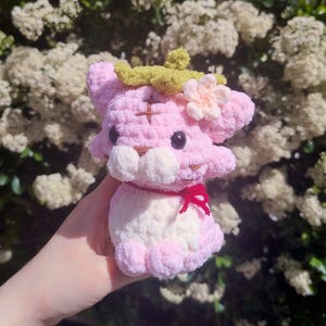 May include: A handmade pink and white crochet tiger plushie with a green leaf and flower accent on its head. The tiger has a red bow around its neck and black eyes. The plushie is held in front of a background of white flowers.