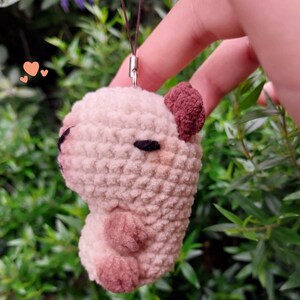 May include: A crocheted capybara charm in shades of beige and brown, with a small loop for attachment. The charm features a textured, plush appearance, with dark eyes and a detailed ear. The background is a blurred view of green foliage.