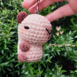 May include: A crocheted plushie of a capybara in light beige and brown. The capybara has a small brown ear, a black eye, and two small brown feet. The plushie is attached to a black string with a silver clasp. The text "@MaykePlushies" is visible.