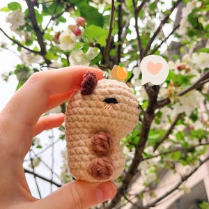 May include: A crocheted keychain in the shape of a capybara. The keychain is beige with brown accents for the ears and feet. A small orange charm and a speech bubble with a heart are attached to the keychain.