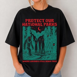 Może przedstawiać: Czarny t-shirt z grafiką w kolorze czerwonym i morskim. Napis głosi PROTECT OUR NATIONAL PARKS oraz WHERE LEGENDS STILL ROAM FREE. Ilustracja przedstawia leśną scenerię z nietoperzem, sasquatchem i innymi mitycznymi stworzeniami.