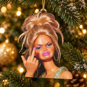 May include: A flat, hanging ornament featuring a doll with blonde hair styled in a high bun, wearing large hoop earrings and a glittery gold top. The doll has a frowning expression, bright pink lipstick, and is holding up a middle finger.