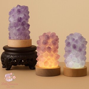 May include: Three amethyst crystal lamps with wooden bases. One lamp is on a black decorative stand. The lamps are various shades of purple and pink, with one illuminated from within, casting a warm glow.