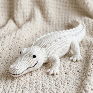 Op de afbeelding: Een wit gehaakt alligator speelgoed met een lachend gezicht en kleine zwarte ogen. Het handgemaakte knuffeldier ligt op een crèmekleurige, getextureerde deken. De alligator heeft een lang lichaam met een getextureerde rug en vier kleine voetjes.
