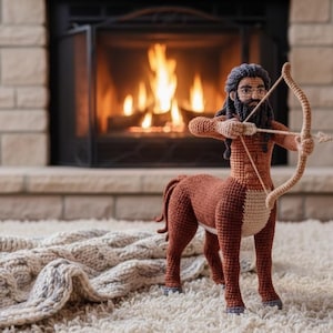 May include: A handmade crochet centaur figurine with a bow and arrow, in shades of brown, tan, and black. The mythical creature stands on a white rug in front of a fireplace with a burning fire. A knitted blanket is in the foreground.