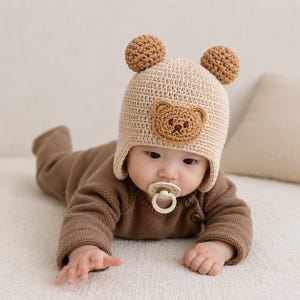 May include: A baby wearing a beige crocheted bear hat with two brown pom-poms and a bear face. The baby is also wearing a brown sweater and a dummy.