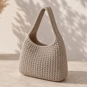 May include: A beige crochet handbag with a rounded shape and a single handle. The bag is made of a textured yarn, and the handle is attached to the top of the bag. The bag is sitting on a light-colored surface.