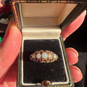 May include: A gold ring with opals and small red gemstones, presented in a black jewellery box. The ring has a filigree design, set with five opals and numerous red stones. The box has a black exterior and a black velvet interior.