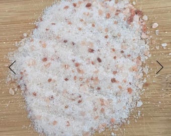 May include: A pile of coarse, pink Himalayan salt crystals on a light brown wooden surface. The salt has a granular texture with varying shades of white and pink, and is scattered across the wood.