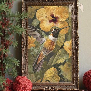 May include: Framed cross-stitch art featuring a hummingbird in flight, surrounded by yellow hibiscus flowers and green leaves. The bird has brown, green, and white feathers. The frame is ornate with a gold finish. The artwork is displayed on a wooden surface.